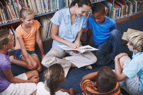 Elementary school teacher and kids sitting in school library, looking at a picture book