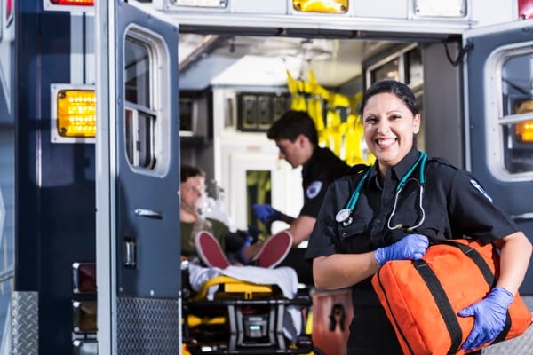 A female first responder stands while another responder tends to a patient in an ambulance.