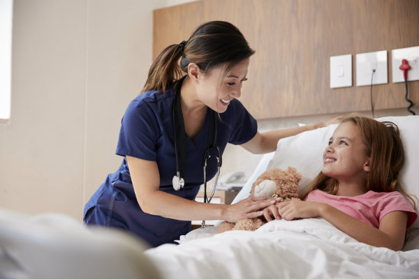 A female nurse visits a girl lying in a hospital bed with a teddy bear
