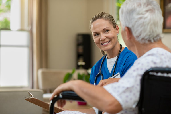 A female nurse with a clipboard sitting down with an elderly patient in a wheelchair.