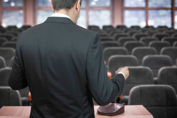 A man stands at a lectern before a large meeting room full of empty chairs.