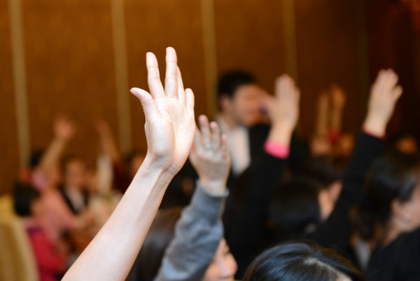 A crowd in a large meeting room raise their hands.